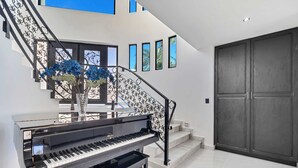 A grand piano near a staircase with a decorative railing, a large dark wooden door, and a wall with small windows. - Marbella Lane Coastal Elegance Villa with Pool (Cabo San Lucas)