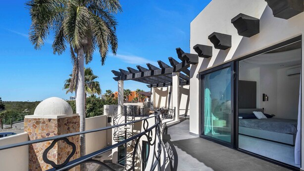A balcony with a glass door leading to a bedroom, a stone wall, and a palm tree. - Marbella Lane Coastal Elegance Villa with Pool (Cabo San Lucas)