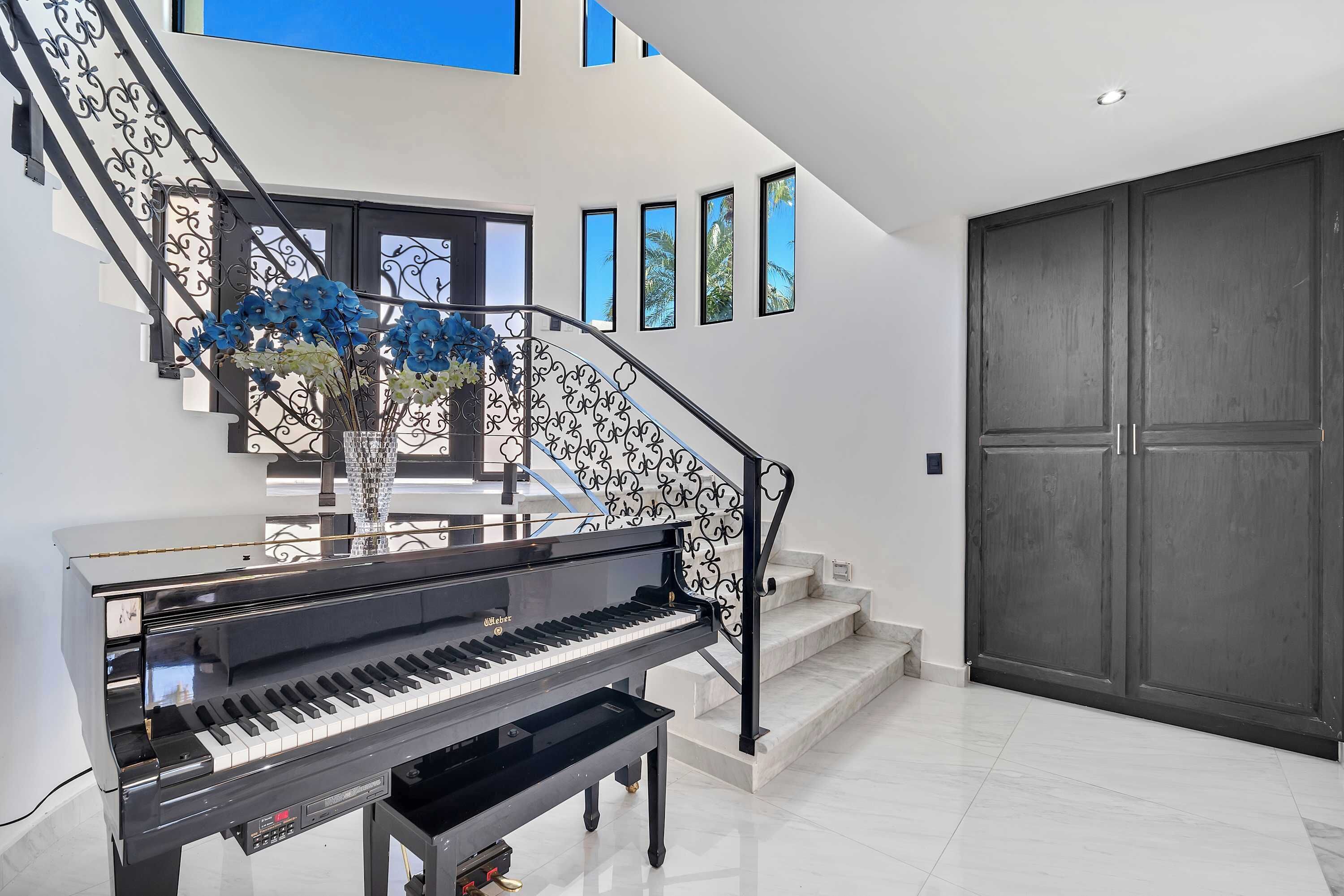 Un piano à queue se trouve près d’un escalier orné d’une rampe décorative, d’une grande porte en bois sombre et d’un mur percé de petites fenêtres.