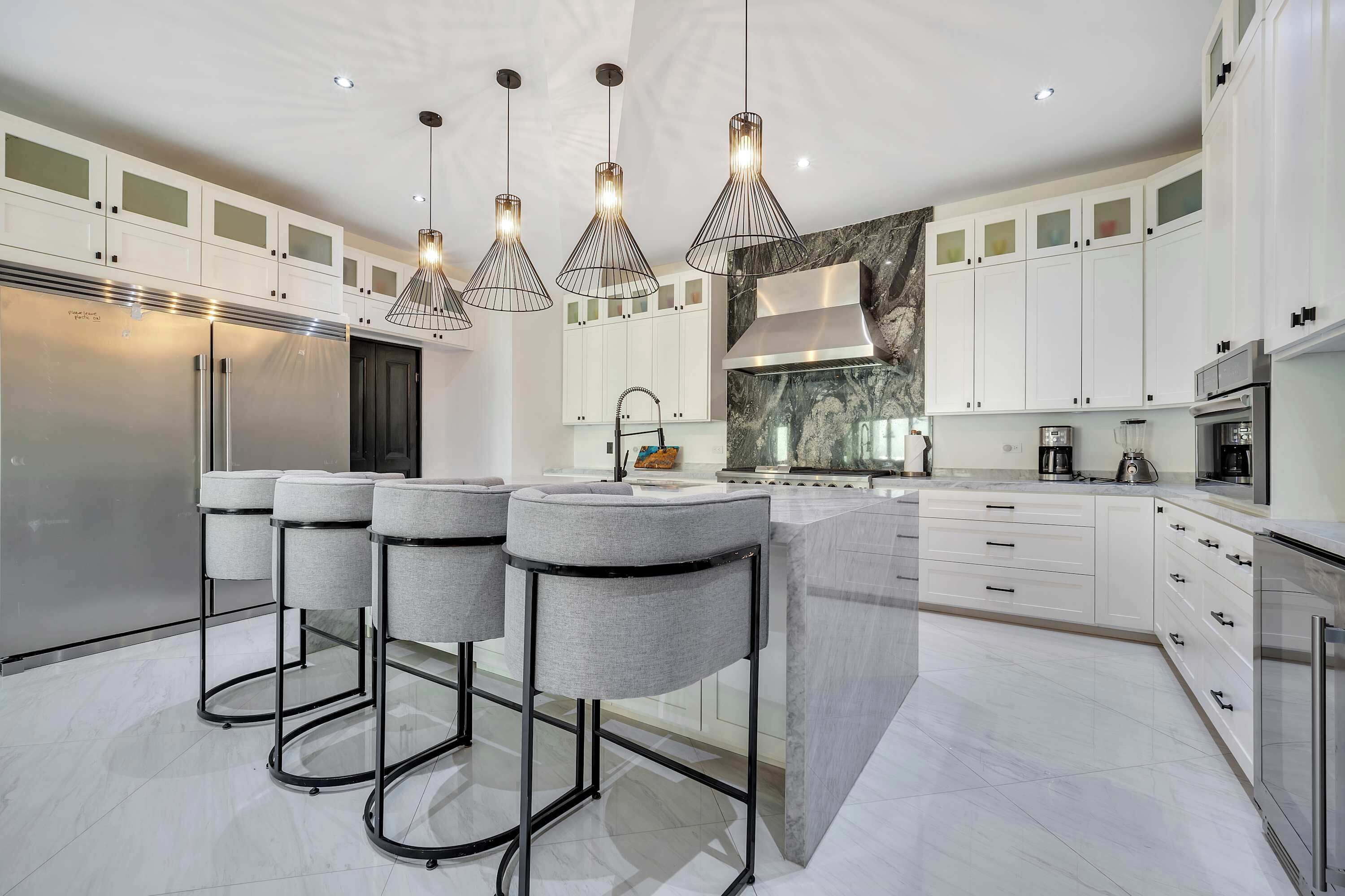 Una cocina moderna con gabinetes blancos, una isla central, taburetes de bar e iluminación colgante.