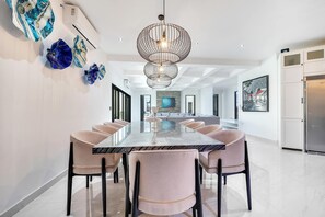 A modern dining area with a marble table and cushioned chairs, a unique chandelier, and a kitchen with a refrigerator. - Marbella Lane Grand 5BR Oasis Villa Getaway (Cabo San Lucas)