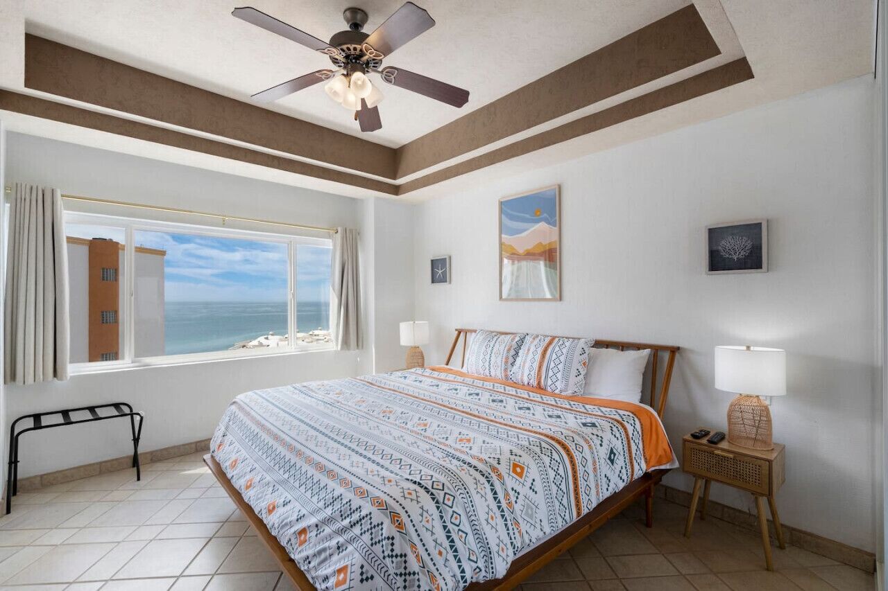 A bedroom with a bed, a ceiling fan, a window with a view of the sea, and a bedside table with a lamp.