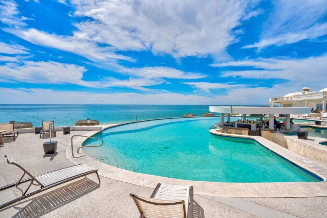 A pool area with lounge chairs, a bar, and a view of the ocean.