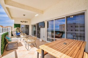En rummelig balkon med et træbord og fletstole, store skydedøre af glas og udsigt over bybilledet.