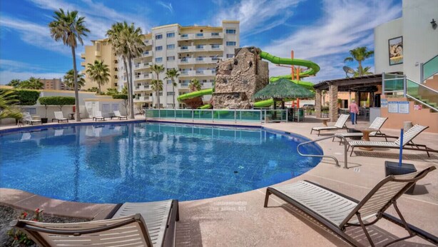 Uma piscina com espreguiçadeiras, um tobogã e um edifício com várias varandas ao fundo.