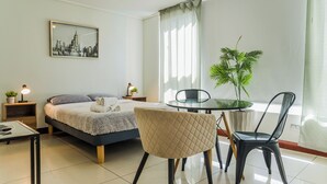 A modern bedroom with a bed, bedside tables, a desk, a chair, a round table, a potted plant, and a cityscape painting on the wall. - Charming Studio in Las Condes  (Las Condes)