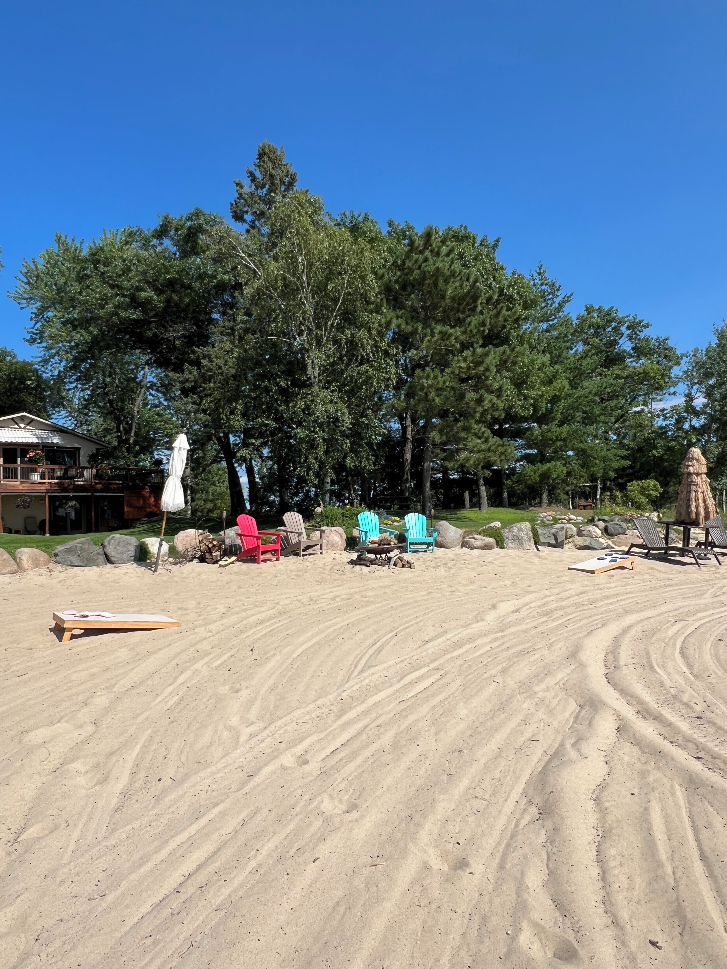 A sandy beach area with lounge chairs, a fire pit, and a large tree in the background.