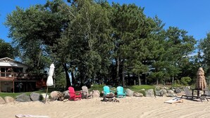 Ein Strandbereich mit Sand, Liegestühlen, einer Feuerstelle und einem großen Baum im Hintergrund.