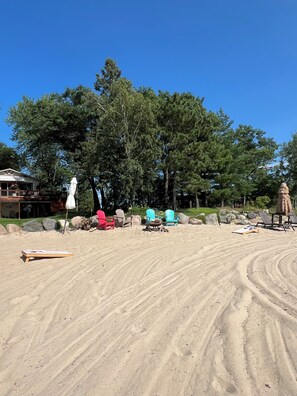 Ein Strandbereich mit Sand, Liegestühlen, einer Feuerstelle und einem großen Baum im Hintergrund.