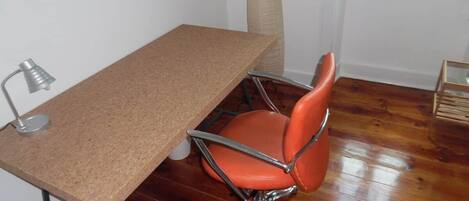 Una habitación con suelo de madera, un escritorio marrón con lámpara, una silla naranja y una ventana con persianas.