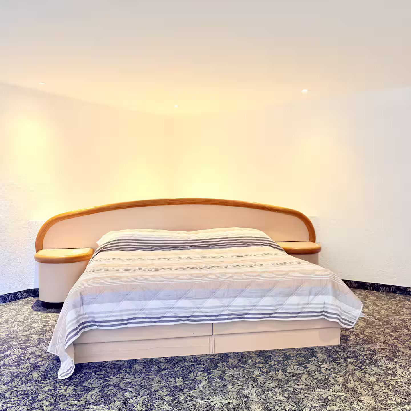 A neatly made bed with a striped bedspread and wooden headboard, positioned against a plain wall with a patterned carpet.