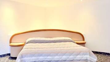 A neatly made bed with a striped bedspread and wooden headboard, positioned against a plain wall with a patterned carpet.