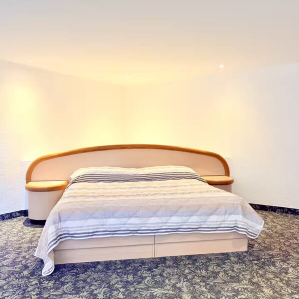 A neatly made bed with a striped bedspread and wooden headboard, positioned against a plain wall with a patterned carpet.