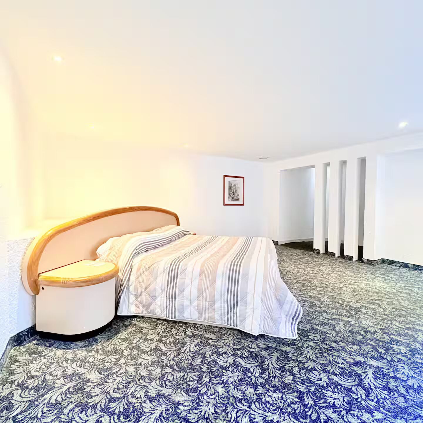 A bedroom with a bed, a bedside table, a framed picture on the wall, and a patterned carpet.