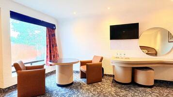 A hotel room with a round table, two armchairs, a sofa, a large mirror, and a television mounted on the wall.