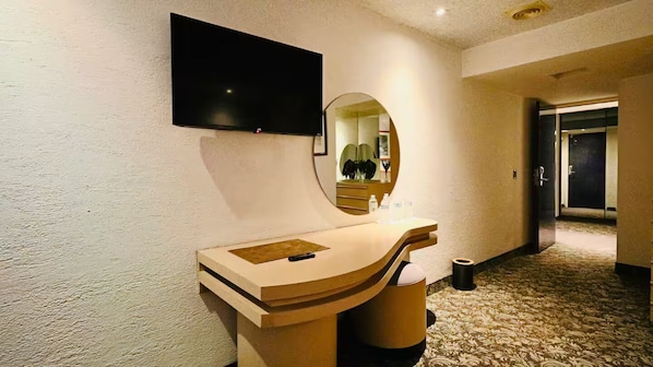 Une chambre d’hôtel dotée d’un téléviseur à écran plat fixé au mur, d’un miroir rond, d’une coiffeuse en bois avec un lavabo et d’un sol recouvert de moquette.