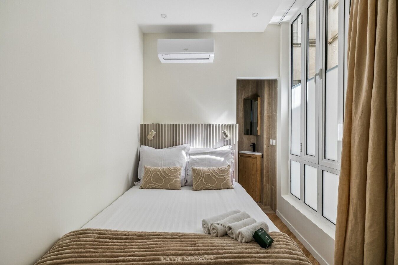 A hotel room with a bed, a window with curtains, a wall-mounted air conditioner, and a wooden cabinet.