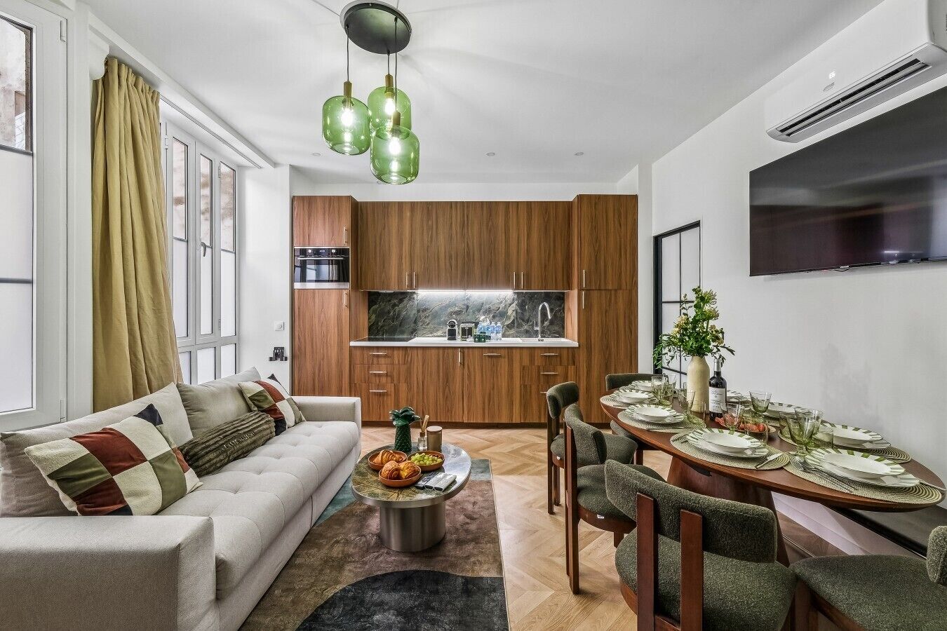A modern living room with a sofa, dining table set for four, and a kitchen with wooden cabinets and a built-in oven.
