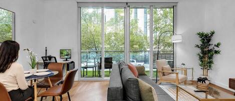 A modern living room with a sofa, chairs, a dining table, and a large window with a view of trees and buildings.