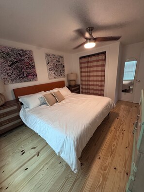A bedroom with a wooden floor, a bed with white bedding, a ceiling fan, and two wall art pieces. - Cozy renovated 2-bedroom/1 bath cottage only 1 block from the beach! (Folly Beach)