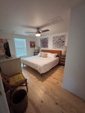 A bedroom with a bed, a desk, a chair, a ceiling fan, a window with blinds, and wall art. - Cottage beside Bert's in the Heart of Folly Beach-one block to the beach & pier! (Folly Beach)