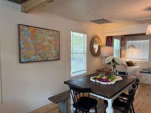 Uma sala de jantar com mesa de madeira, cadeiras, um banco, uma pintura abstrata colorida, um espelho e uma janela com persianas.