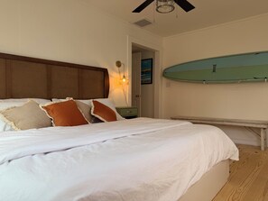 Quarto com cama, almofadas, mesa de cabeceira, prancha de surf na parede e ventilador de teto.
