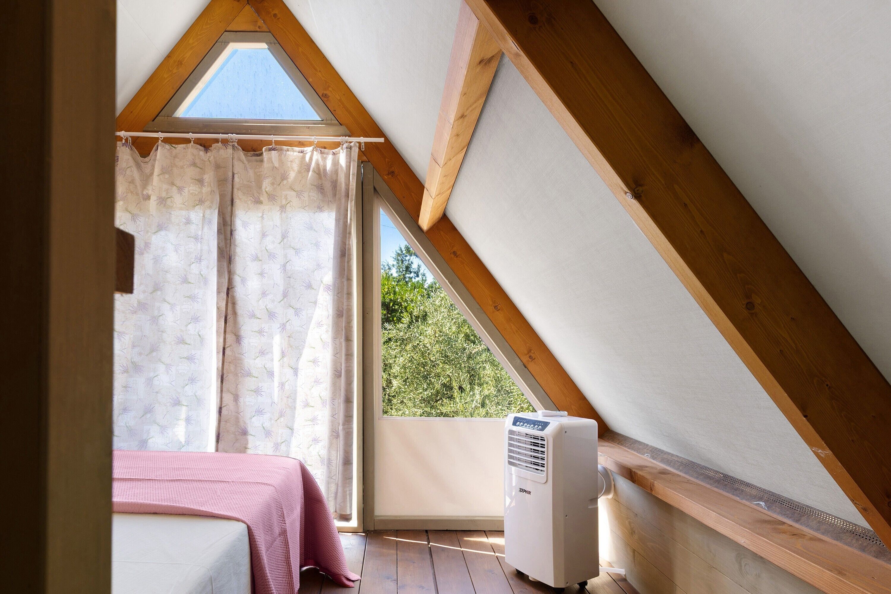 A room with a sloped ceiling, a bed with a pink blanket, a window with a curtain, and a white air conditioner.