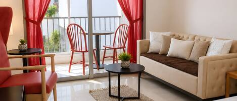 A hotel room with a bed, a sofa, two red chairs, a small table with a plant, and a balcony with red curtains.
