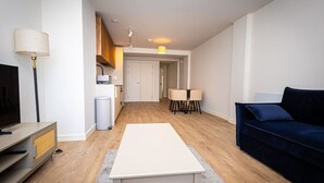 Uma sala moderna com televisão de tela plana, sofá azul escuro, mesa de centro branca e um espaço de refeições com duas cadeiras e uma mesa pequena.