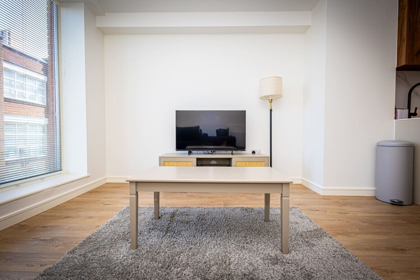 A modern living room with a flat-screen TV on a white stand, a grey carpet, and a lamp on the right.