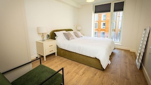 Quarto com cama, mesas de cabeceira, uma cadeira, janela com vista para edifícios e radiador.
