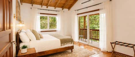 A bedroom with a wooden ceiling, a bed with white bedding, a small bedside table with a lamp, and a view of greenery through the windows.