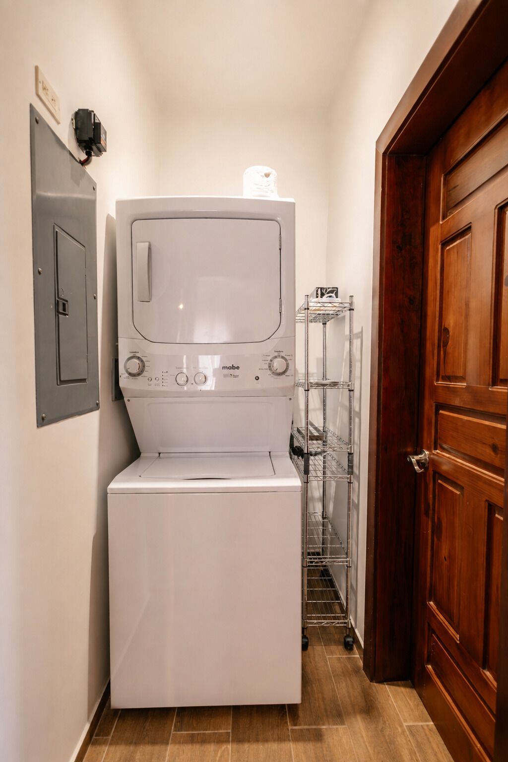 Uma máquina de lavar e secar compacta, branca e fixada na parede, num corredor estreito com uma porta de madeira e uma estante de metal na parede.