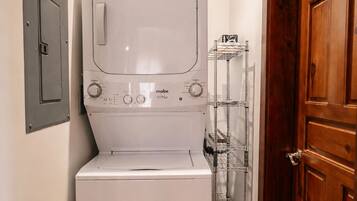 Uma máquina de lavar e secar compacta, branca e fixada na parede, num corredor estreito com uma porta de madeira e uma estante de metal na parede.