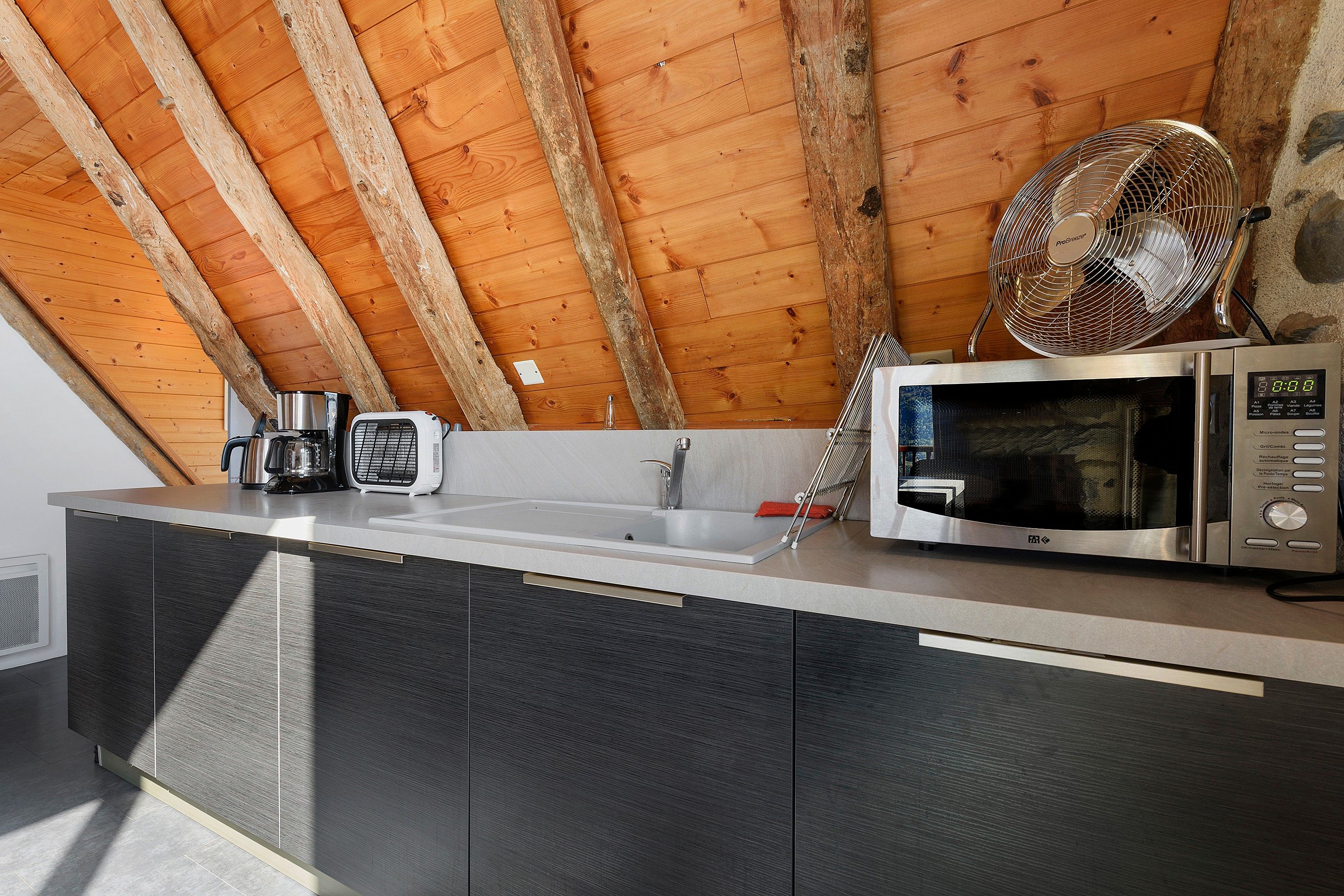 A kitchen with a wooden ceiling, a sink, a microwave, and a kettle on the counter.