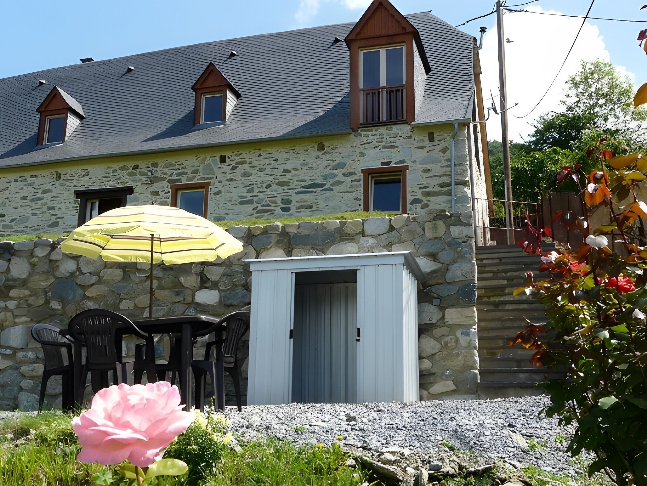Une maison en pierre avec un toit en pente, un petit abri attenant, un espace extérieur aménagé pour s’asseoir et une fleur rose.