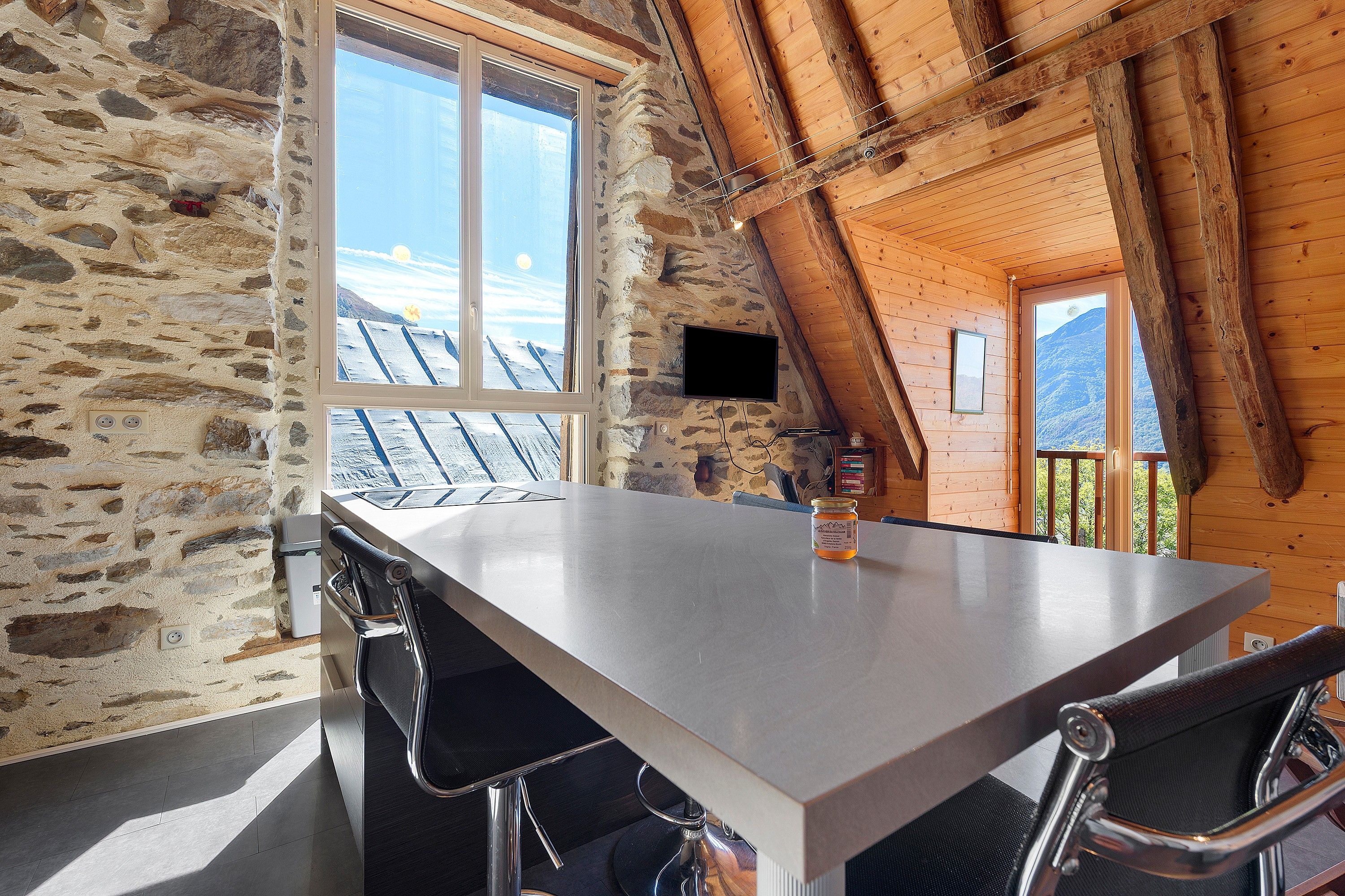 Un mur de pierre, une fenêtre donnant sur des toits enneigés, une table et des chaises dans une pièce aux poutres apparentes.