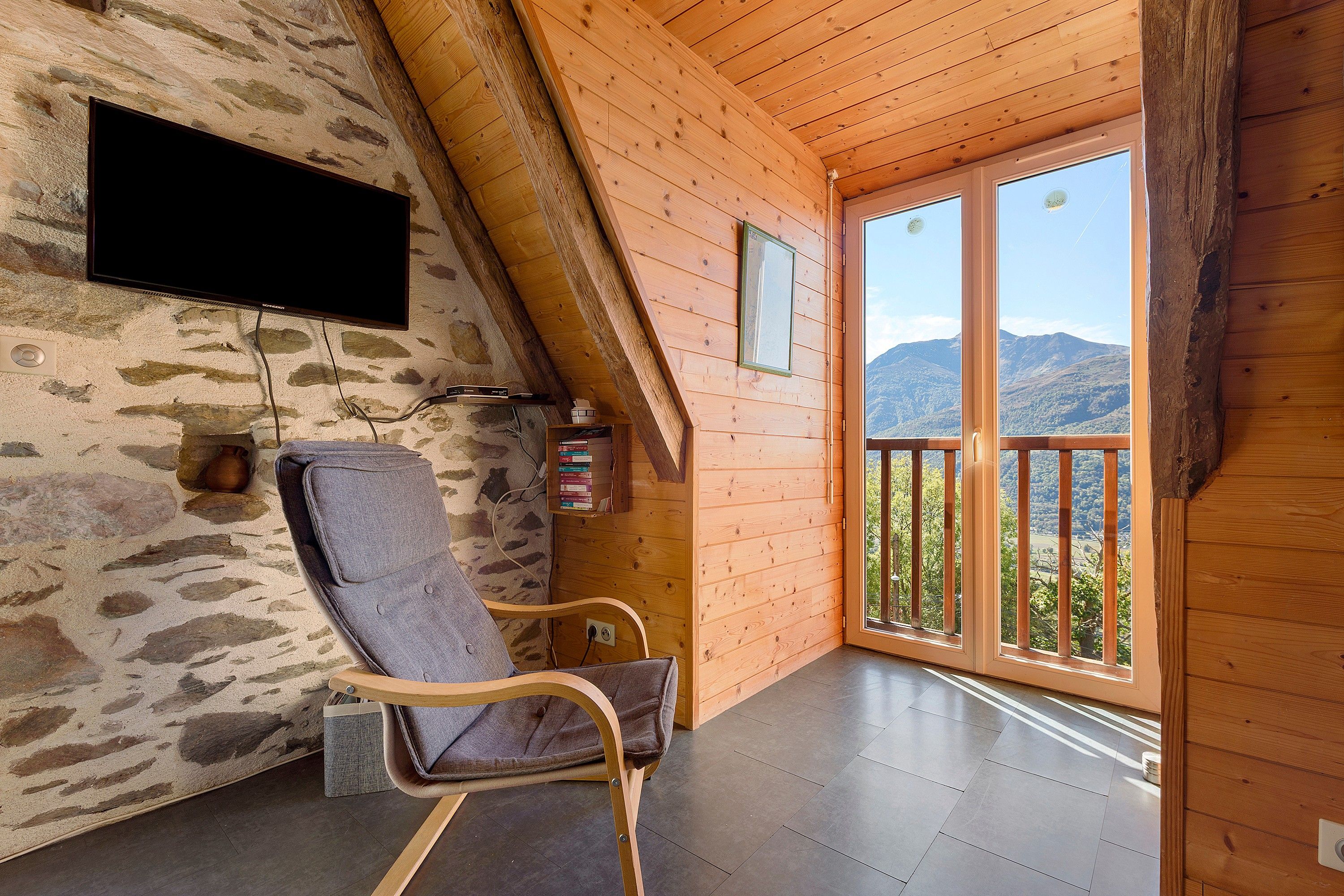 A cozy room with a stone wall, a flat-screen TV, a wooden chair, and a large window with a mountain view.
