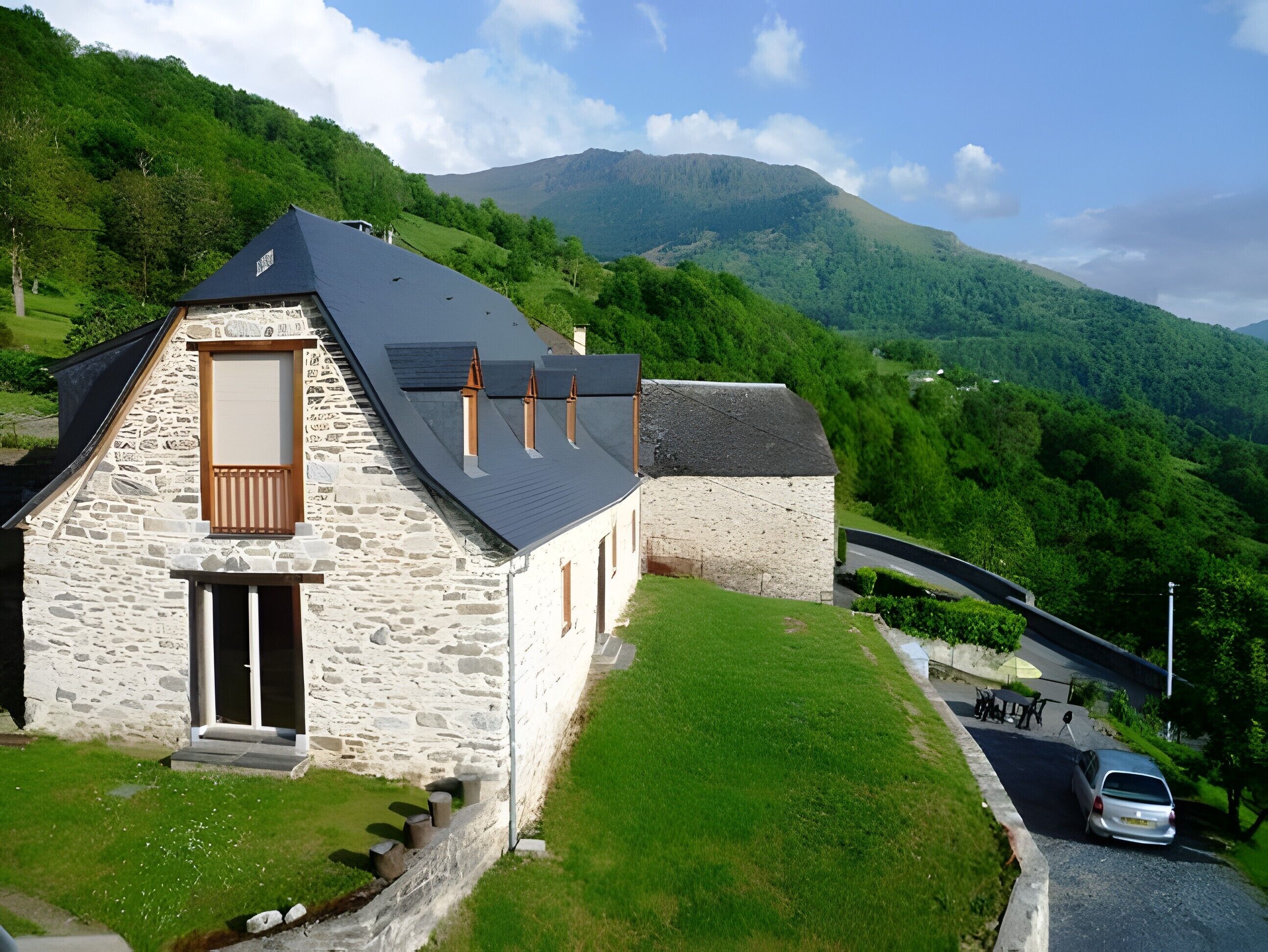 Una casa de piedra con techo inclinado, un pequeño porche y vistas a un valle verde y montañas.