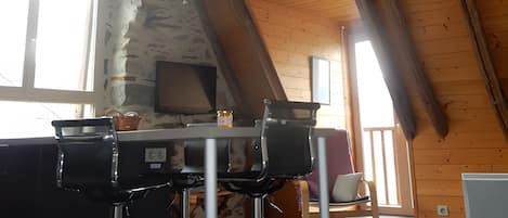 Un interior rústico con techo inclinado, pared de piedra, mesa de madera y dos taburetes de bar.