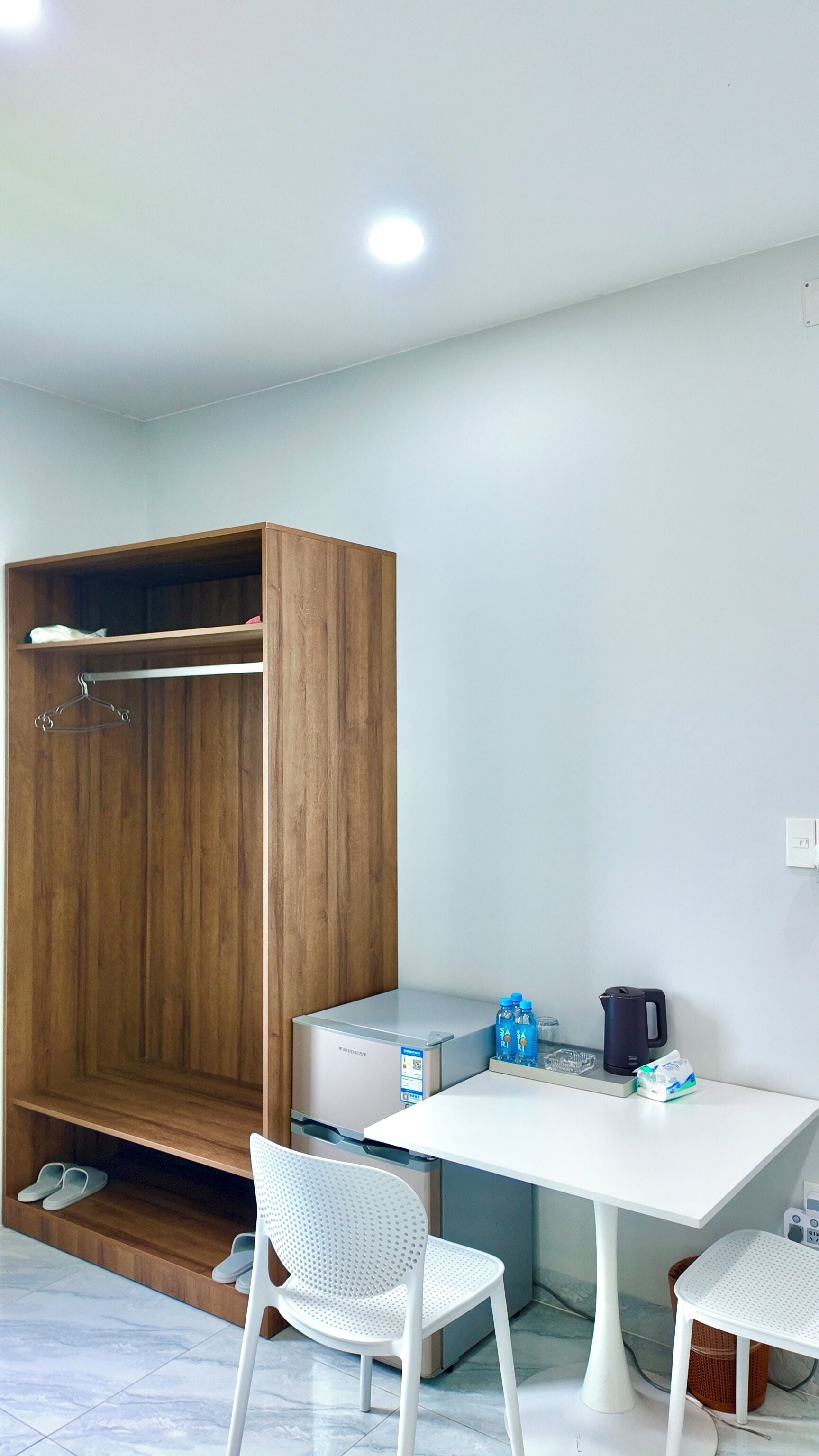 A room with a wooden wardrobe, a white table with a kettle and water bottles, and two white chairs.