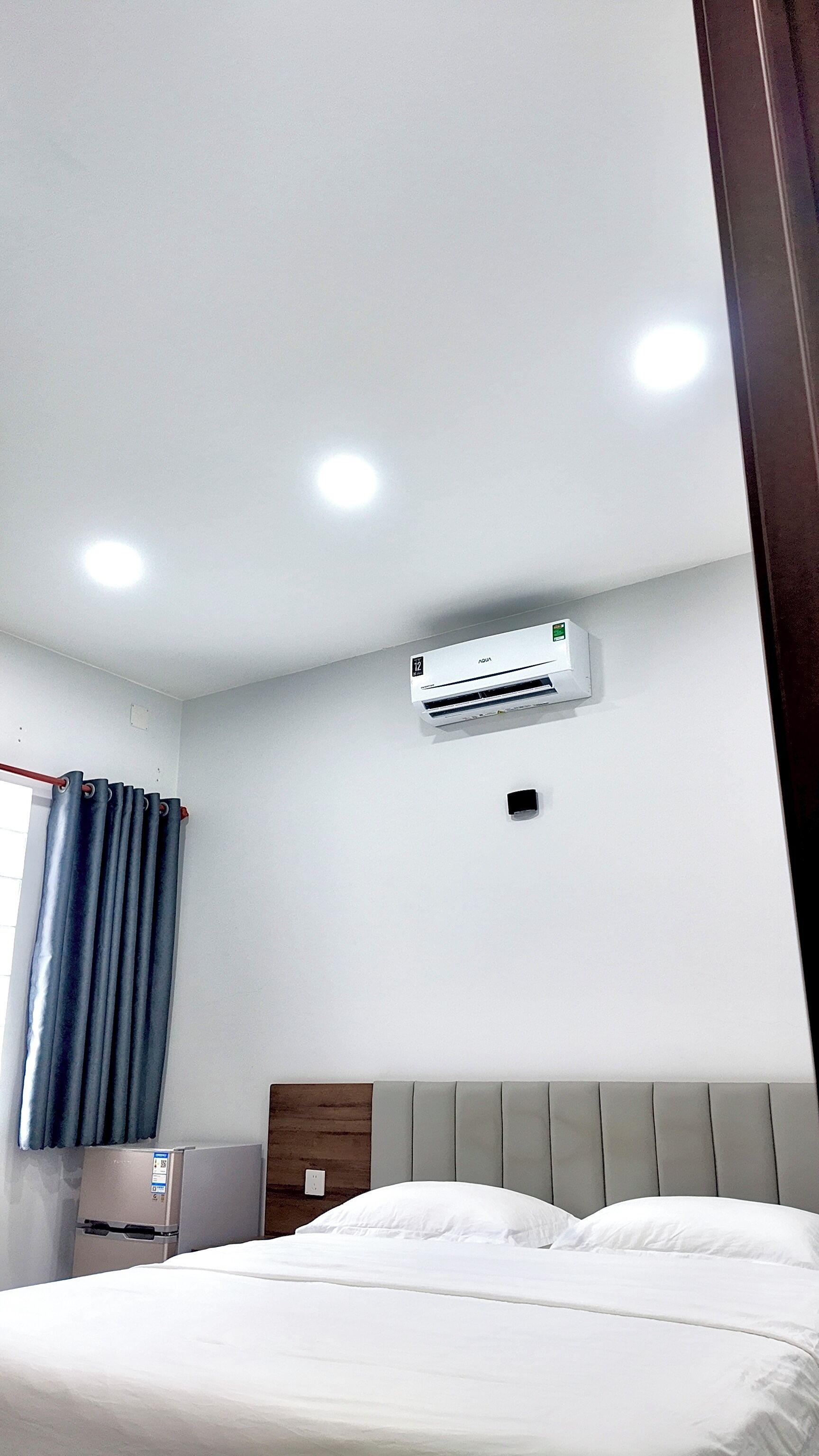 A hotel room with a bed, a wall-mounted air conditioner, a window with curtains, and a small table with a refrigerator.