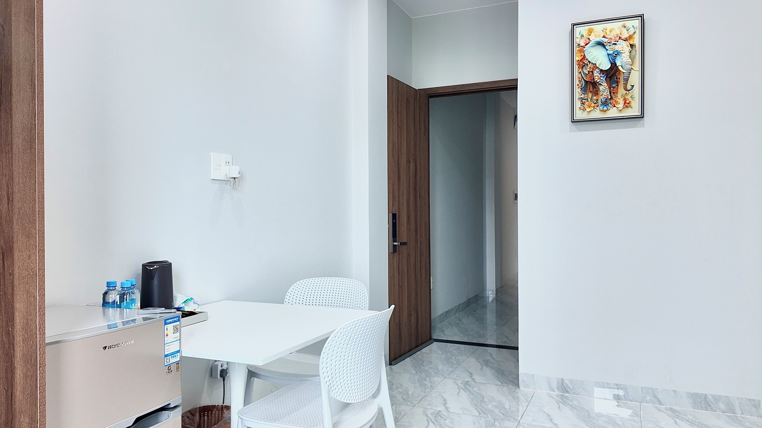 A small, clean room with a white table, two white chairs, a wooden door, and a framed picture on the wall.