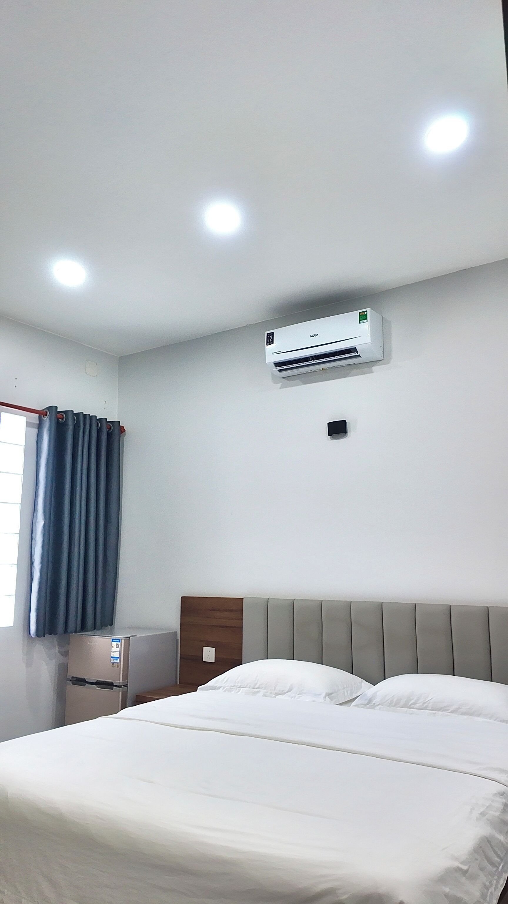 A hotel room with a bed, a wall-mounted air conditioner, a window with curtains, and a small refrigerator.