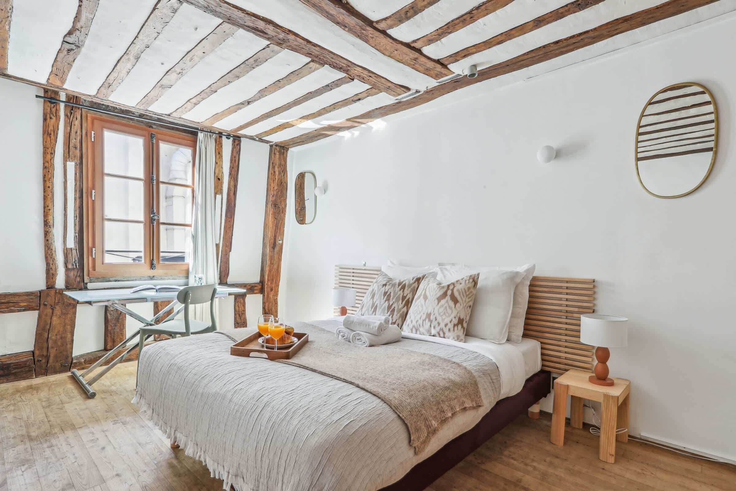 A bedroom with a wooden ceiling, a bed with a headboard, a bedside table with a lamp, and a window with curtains.
