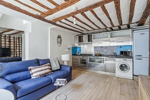 A modern living room with a blue sofa, a washing machine, and a kitchen with stainless steel appliances. - Bright 1BR - Walk to Louvre - Opera (Paris)