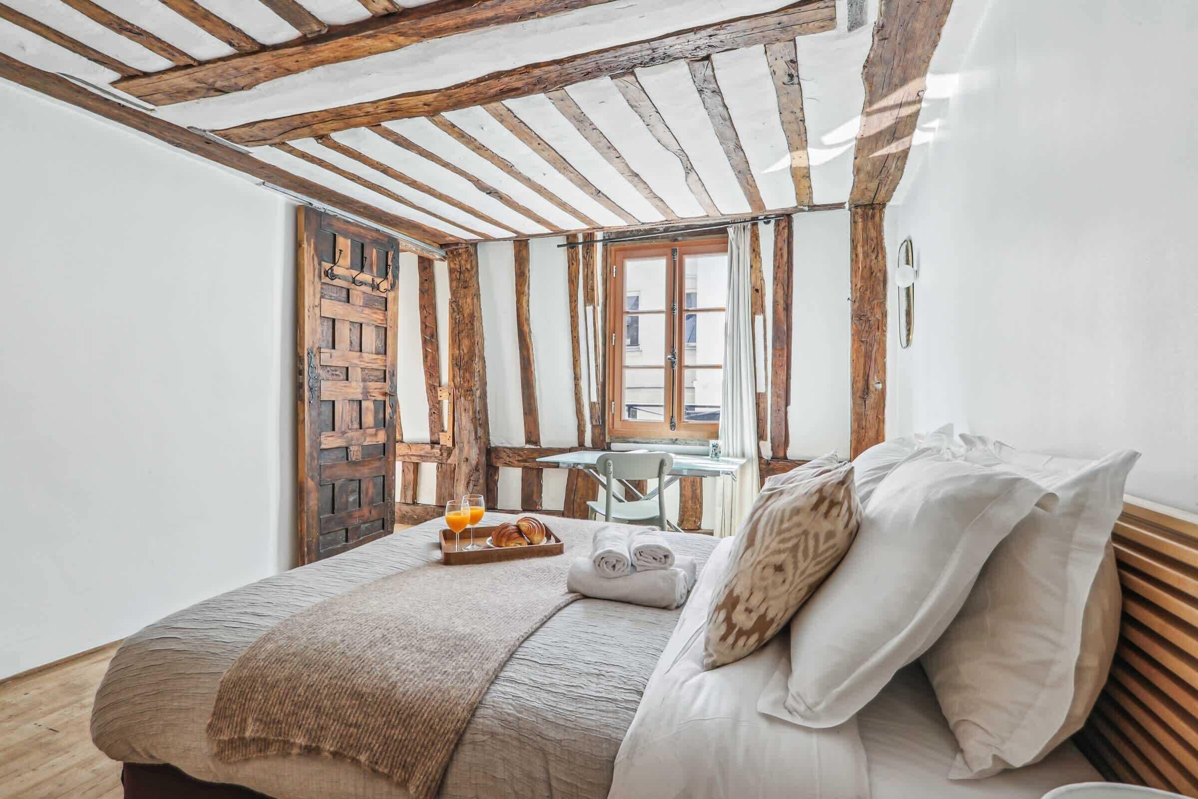 A cozy bedroom with a wooden ceiling, a bed with white linens, a small table with breakfast, and a window with a view.