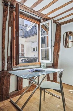 A room with a glass table and chair, wooden beams, a large window, and a view of buildings outside. - Bright 1BR - Walk to Louvre - Opera (Paris)
