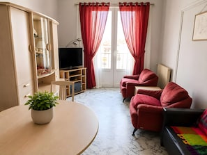 Ein Wohnzimmer mit Fernseher, roten Vorhängen, einer Couch, einem Esstisch mit Pflanze und einem Heizkörper.